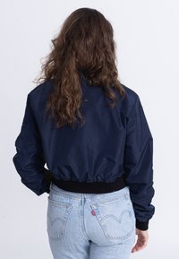 Giacca bomber navy con design cropped, orlo elastico e polsini neri, abbinata a jeans Levi's azzurri chiari con dettagli di cucitura in vista.