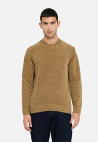 Maglione a maglia marrone con texture, colletto rotondo e polsini a coste. Indossato con pantaloni in denim scuro.
