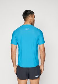 Camiseta atlética azul de manga curta com decote redondo, apresentando um logótipo na parte superior das costas, combinada com calções pretos. Tecido de textura suave.