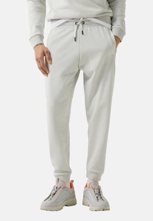 Man draagt lichtgrijze joggerbroek met trekkoorden in de taille en bijpassende sneakers met oranje accenten, staat met zijn handen in zijn zakken.