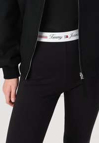 Črna bomberska jakna s srebrnim zadrgo, kombinirana s črnimi pajkicami, ki imajo bel pas z rdečimi in modrimi detajli, blagovna znamka "Tommy Jeans."