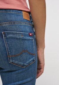 Jeans de mezclilla azul con un ajuste clásico, que cuentan con un bolsillo trasero con etiqueta roja y un parche de cuero marrón, mostrando detalles de costura.