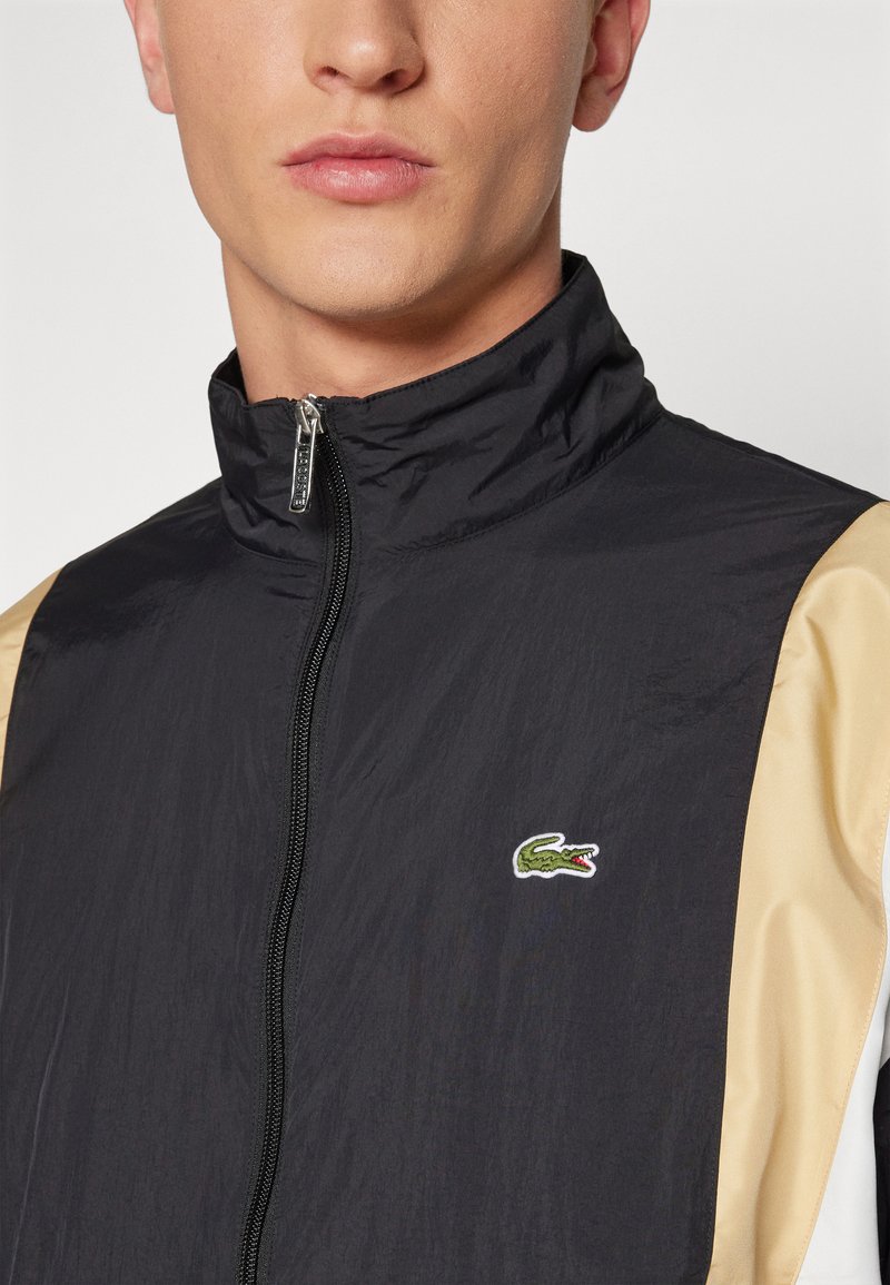 Veste légère noire avec un col haut, des manches beige contrastantes et un petit logo de crocodile vert sur la poitrine. Fermeture éclair à l'avant.