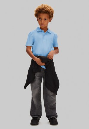 Junge mit lockigem Haar, der ein hellblaues Polohemd, graue Jeans, schwarze Schuhe trägt und eine schwarze Jacke um die Taille hält.