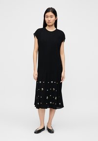 Robe noire plissée à manches courtes, ornée d'une broderie florale colorée sur l'ourlet. Le tissu a une texture lisse. Portée avec des chaussures plates noires.