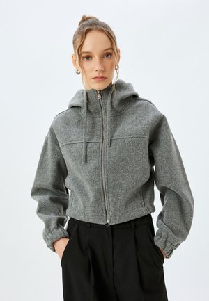 Grau, Zip-Jacke aus strukturiertem Stoff, mit Kapuze, elastischen Bündchen und cropped Design, kombiniert mit schwarzen Hosen.
