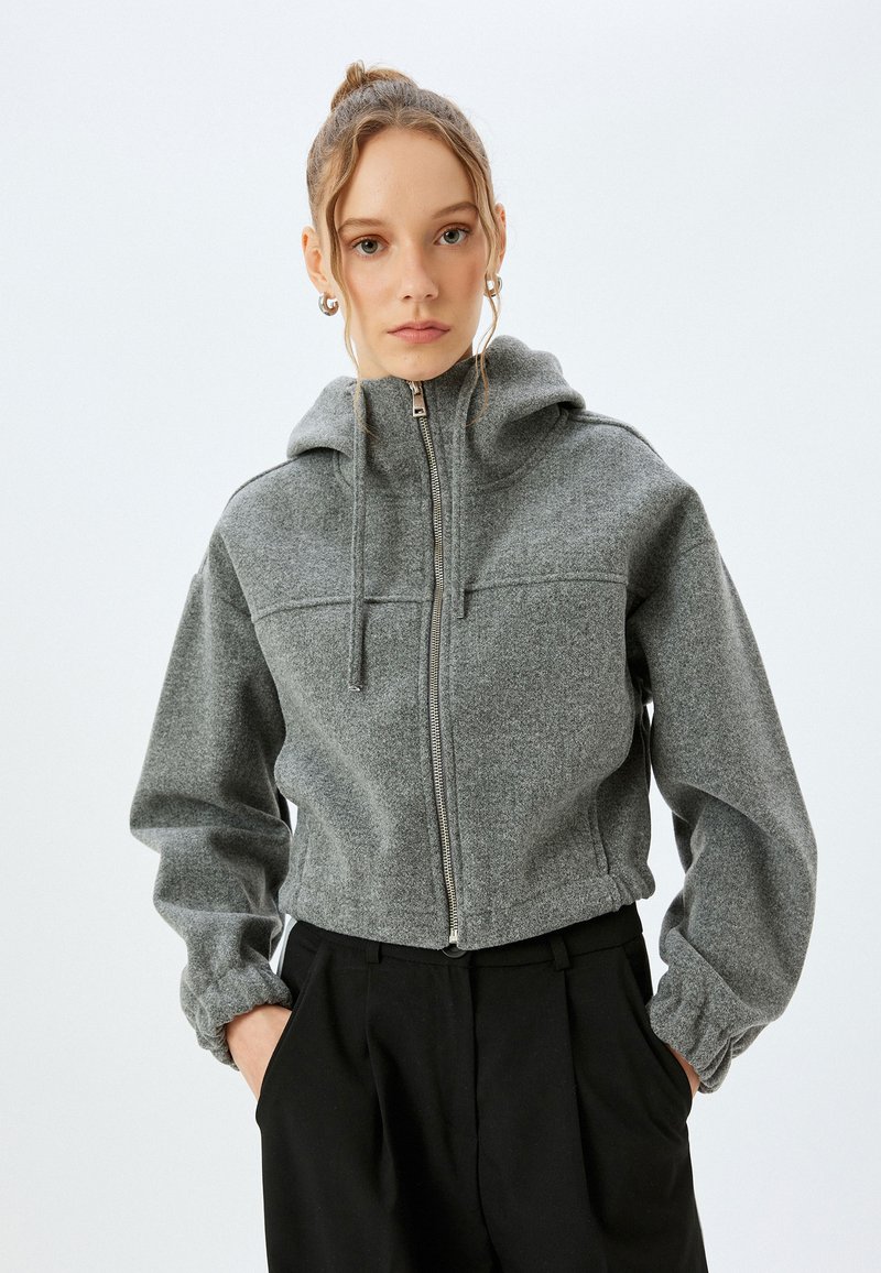 Giacca grigia con zip realizzata in tessuto testurizzato, dotata di cappuccio, polsini elasticizzati e un design corto, abbinata a pantaloni neri.