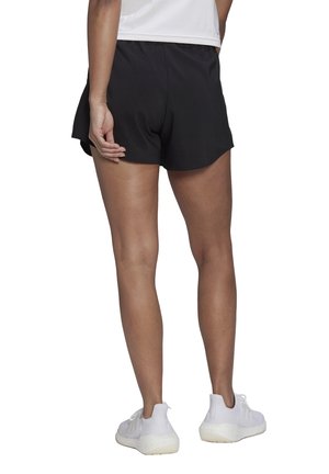 Person iført sorte træningsshorts, en hvid top og hvide sneakers, der står med ryggen mod kameraet på en hvid baggrund.