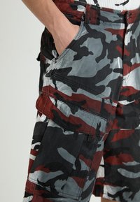 Bermudas cargo de camuflaje con una mezcla de patrones en negro, gris y rojo, múltiples bolsillos y una textura suave. Detalles de herrajes metálicos visibles.