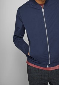 Man som bär en marinblå zip-up jacka med en röd skjorta under och grå rutiga byxor, hand i vänster jackficka, mot en grå bakgrund.