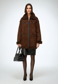 Chaqueta oversize de ante marrón con cuello de borreguillo, ribetes negros a contraste, cremallera frontal y puños acanalados, combinada con falda de cuero negra de talle alto.