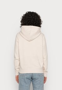 Persona con capelli ricci scuri che indossa una semplice felpa con cappuccio beige chiaro e jeans azzurri chiari, vista di spalle.