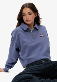 Sudadera de forro polar morada con collar y media cremallera, que presenta un pequeño parche con una bandera americana. Combinada con pantalones oscuros de canalé.
