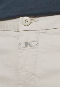 Beiga bomullbyxor med knappstängning och etikett som säger "International Closed Clothing." Slät textur med subtila sömnadsdetaljer.