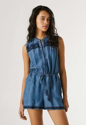 Frau mit langen dunklen Haaren, die einen ärmellosen blauen Denim-Overall mit schwarzer Stickerei und einem Taillen-Kordelzug trägt, steht vor einem schlichten Hintergrund.