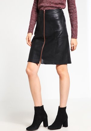 Femme portant une jupe en cuir noire avec une fermeture éclair avant, un pull bordeaux et des bottines noires à talons, debout devant un fond blanc.