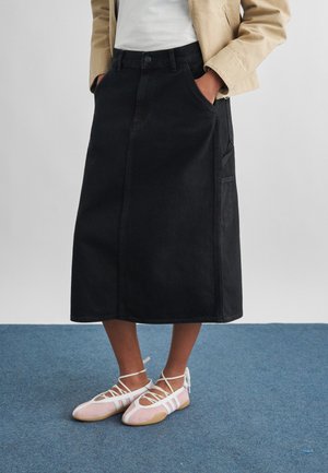 Persoană îmbrăcată cu o geacă bej, fustă midi neagră cu buzunare și balerini roz cu bretele albe, stând pe o mochetă albastră.