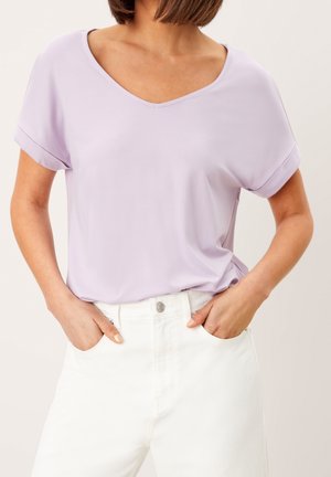 Nainen, jolla on vaaleanvioletti lyhythihainen V-pääntiellä varustettu paita ja joka on työnnetty korkeavyötäröisten valkoisten housujen sisään, kädet taskuissa.