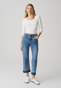 Weißes Langarmoberteil kombiniert mit hochgeschnittenen, hellblauen Jeans im Denim-Stil, die cropped und fransige Säume sowie einen lässigen Schnitt aufweisen. Weiße Sneakers runden das Outfit ab.