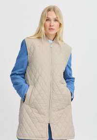Beige gesteppte Weste mit blauen Denimärmeln, ausgestattet mit einem Frontreißverschluss und Seitentaschen, die einen kontrastierenden Farb- und Textureneffekt bieten.