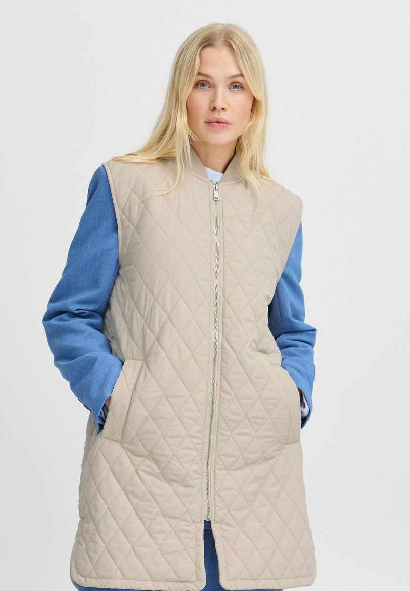 Beige gesteppte Weste mit blauen Denimärmeln, ausgestattet mit einem Frontreißverschluss und Seitentaschen, die einen kontrastierenden Farb- und Textureneffekt bieten.