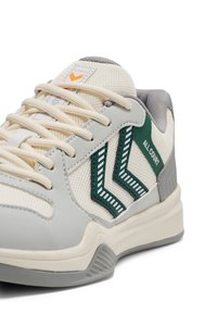 Close-up van een beige en grijze sneaker met groene chevronstrepen en de tekst "ALL COURT" aan de zijkant, met crèmekleurige veters.