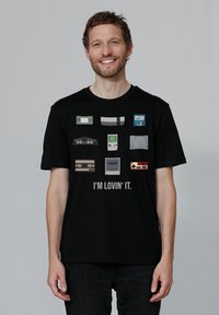 T-shirt noir présentant des arts en pixel de consoles de jeux vintage et de manettes, avec le texte "J'AIME ÇA." sur le devant.