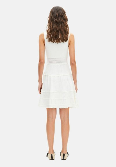 Mujer con cabello castaño ondulado, vestida con un vestido blanco sin mangas con detalles de encaje, de pie y de espaldas, combinando con zapatos beige y negros.
