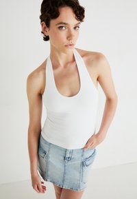 Top alb cu gât halter și decolteu rotunjit, asociat cu o fustă mini din denim bleu deschis, care are două buzunare frontale și o finisare decolorată.