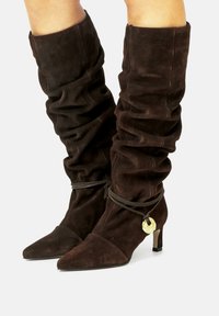 Botas altas de ante marrón con textura recogida, punta afilada y un tacón bajo. Presentan un acento decorativo de hardware dorado y un cierre atado.