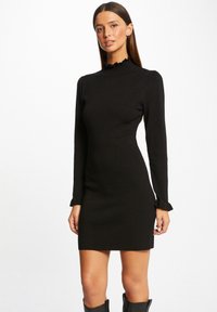 Vestido negro de punto con un alto cuello rizado, mangas largas y puños acampanados. Diseño elegante y ceñido. Textura suave, longitud hasta medio muslo.