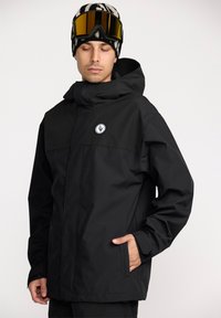 Veste imperméable noire avec capuche, dotée d'un emblème sur le devant et de poches latérales. Texture lisse, design minimaliste, adaptée à un usage en extérieur.