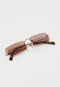 Lunettes de soleil rectangulaires brunes avec un pont doré, des plaquettes de nez transparentes et des branches en écaille de tortue. Verres légèrement teintés pour une protection solaire.