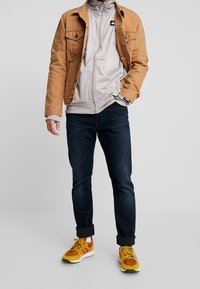 Tan denimjakke over en lys grå zip-up jakke, parret med mørke jeans og multicolore sneakers med gule, orange og hvide accenter.