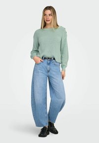 Femme portant un pull en tricot vert menthe avec des détails à boutons, un jean large bleu clair, une ceinture noire et des chaussures noires massives sur un fond uni.