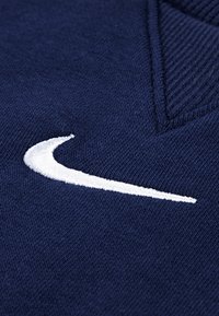 Marineblå strikket genser med V-hals, med en fremtredende hvit brodert Nike swoosh-logo på brystet.