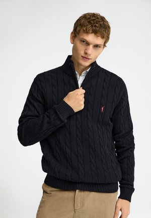 Junger Mann schließt den Reißverschluss an einem schwarzen, grob gestrickten Pullover mit beiger Hose vor einem weißen Hintergrund.