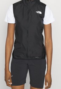 Svart ärmlös väst med hög krage, dragkedja framtill och logodetalj. Bärs över en vit kortärmad skjorta, i kombination med svarta shorts.