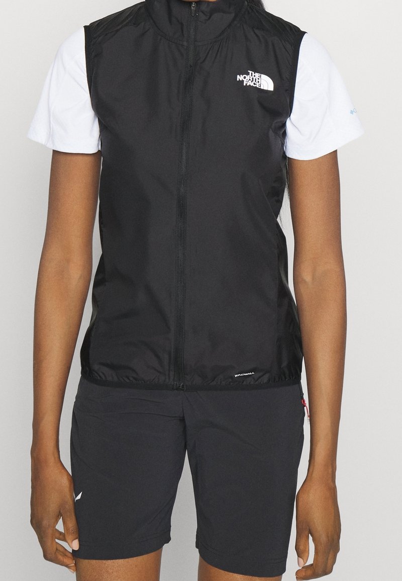 Svart ärmlös väst med hög krage, dragkedja framtill och logodetalj. Bärs över en vit kortärmad skjorta, i kombination med svarta shorts.