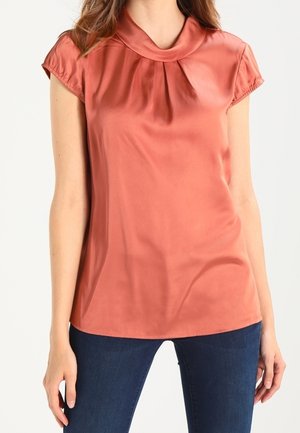 Kvinde iført en kortærmet, satinlignende bluse i støvet rosa med rund krave, kombineret med mørkeblå jeans, står med armene afslappet ned langs siden.
