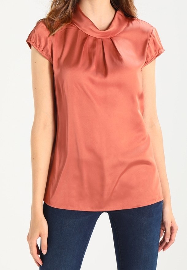 Femme portant un chemisier à manches courtes, satiné, rose poussiéreux avec un col rond, associé à un jean bleu foncé, debout, les bras détendus.