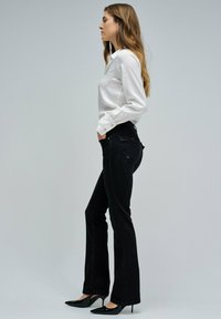 Mujer de pie de lado, vistiendo una blusa blanca, jeans ajustados de campana negros y zapatos de tacón alto negros, sobre un fondo gris liso.