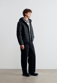 Νεαρός άνδρας όρθιος, φοράει μαύρο μπουφάν, καρό πουκάμισο, μαύρο παντελόνι και μαύρα παπούτσια, μπροστά σε απλό φωτεινό φόντο.