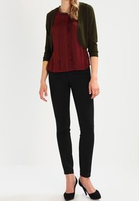 Bordowa bluzka z zakładkami, w połączeniu z ciemnozielonym kardiganem, czarnymi dopasowanymi spodniami i czarnymi szpilkami. Gładka tekstura i dopasowany krój.