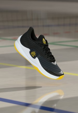 Zwart en geel sportschoen met witte zool zwevend boven een gymzaalvloer gemarkeerd met gekleurde lijnen.