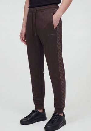 Personne portant un pantalon de jogging marron foncé avec une bande logo latérale et des baskets noires, debout devant un fond clair uni.