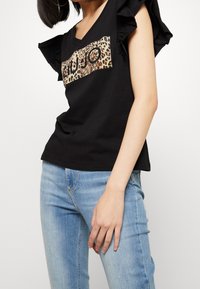 Zwarte T-shirt met korte mouwen, gekenmerkt door ruches op de schouders, een logo patch met luipaardprint en een V-hals ontwerp, in combinatie met lichtblauwe jeans.