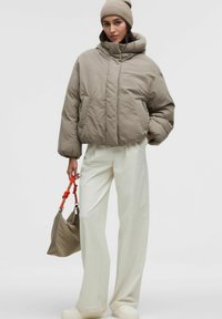 Puchová bunda v taupe farbe s vysokým golierom a prešívanou textúrou, spárovaná s bielymi širokými nohavicami a svetlohnedou taškou s oranžovými akcentami.