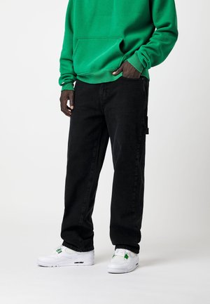 Personne portant un sweat-shirt vert, un pantalon noir avec une boucle latérale et des baskets blanches avec des détails verts, debout devant un fond blanc uni.