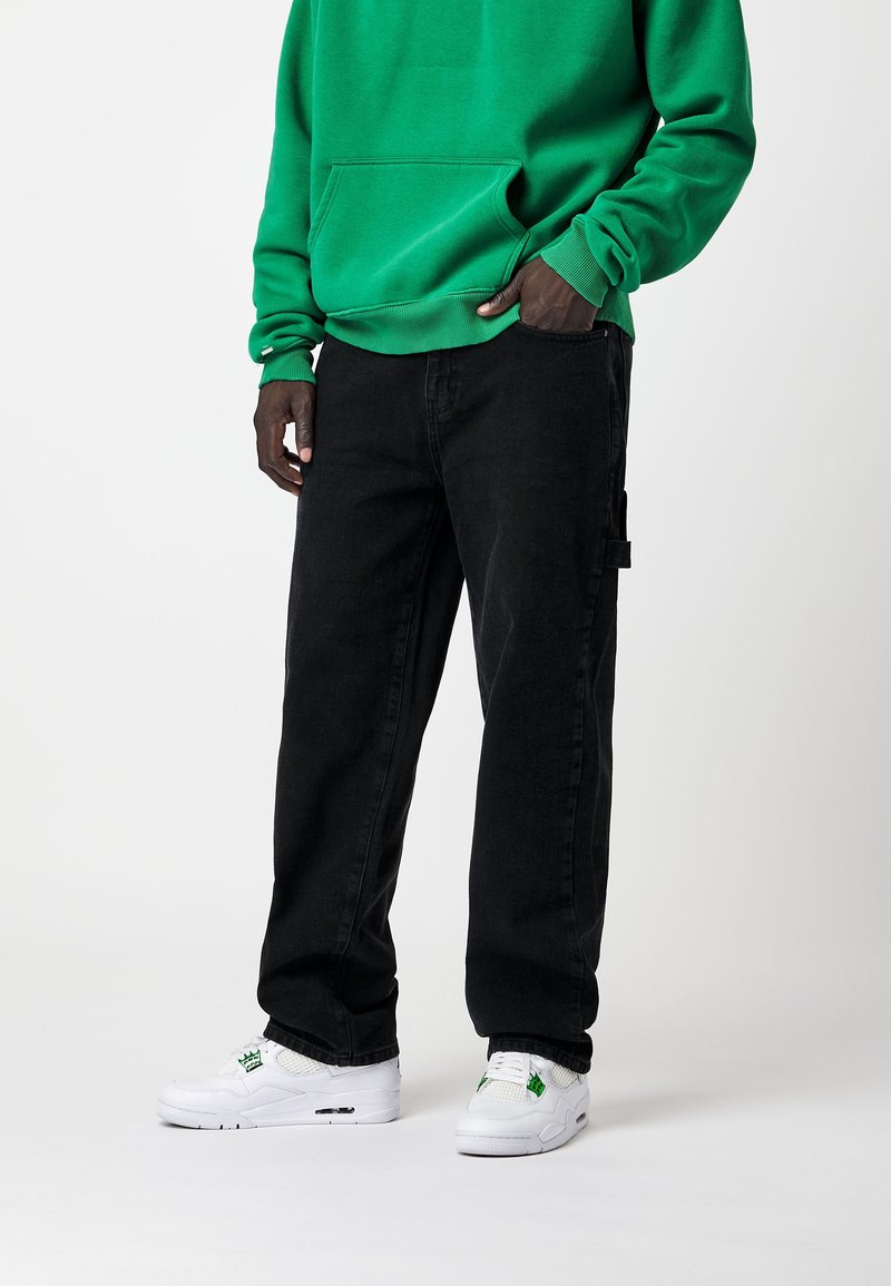 Personne portant un sweat-shirt vert, un pantalon noir avec une boucle latérale et des baskets blanches avec des détails verts, debout devant un fond blanc uni.
